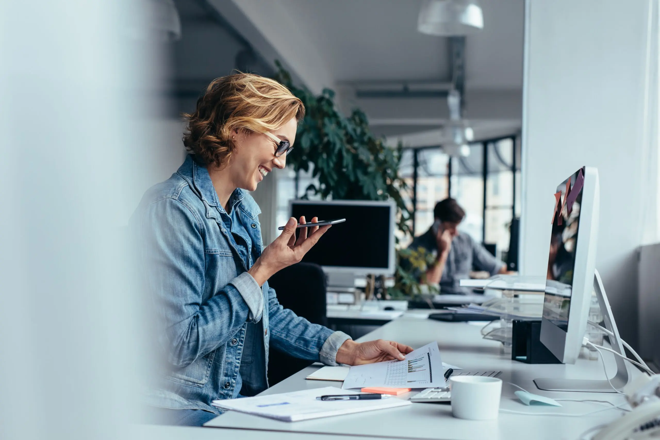 Businesswoman talking on smartphone and looking at documents. Young female executive sitting in front of pc.